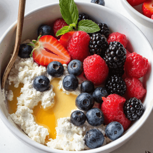 A protein-packed cottage cheese berry bowl topped with fresh strawberries, blueberries, raspberries, blackberries, and a drizzle of honey, served in a white bowl.