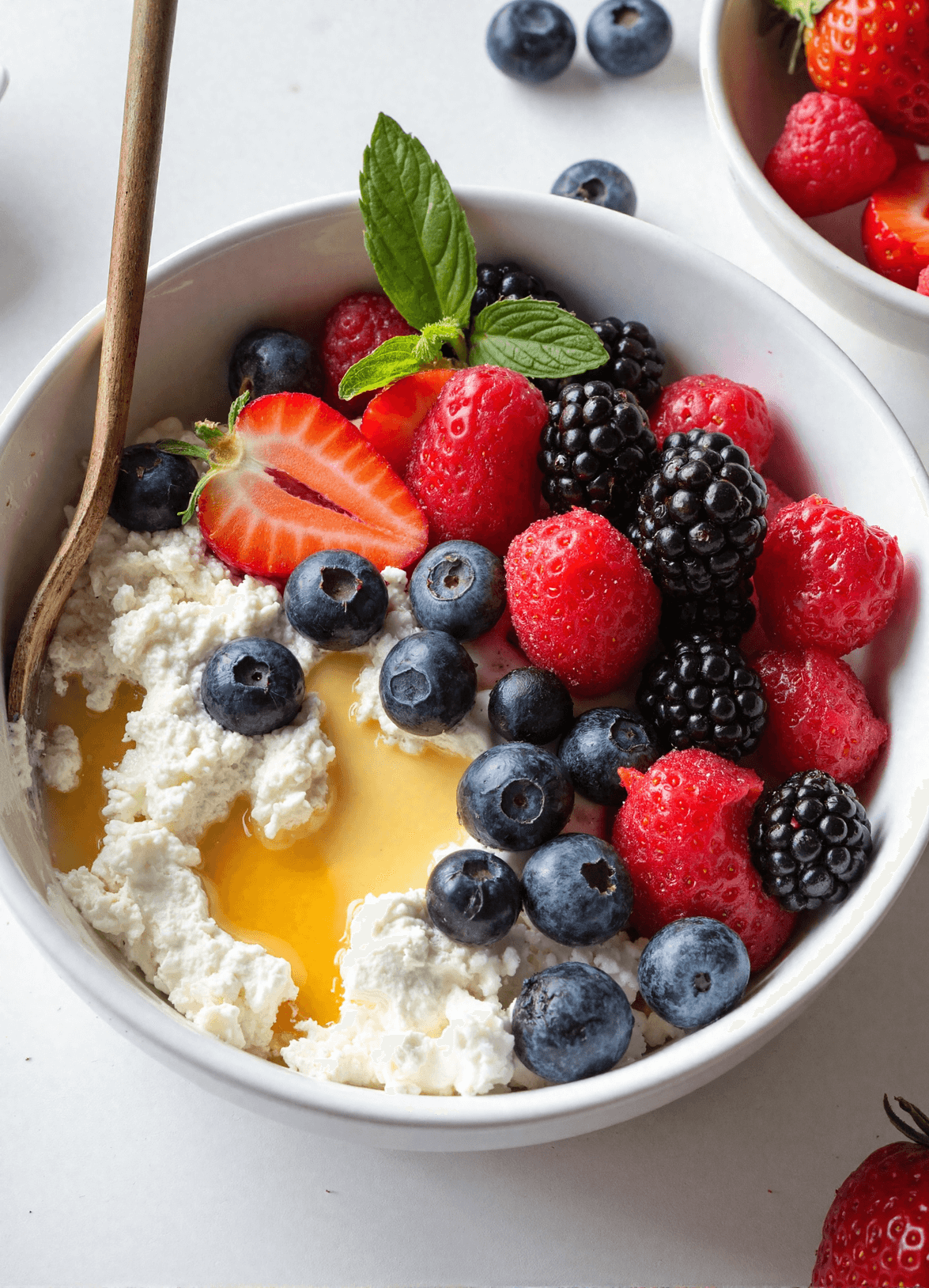 A protein-packed cottage cheese berry bowl topped with fresh strawberries, blueberries, raspberries, blackberries, and a drizzle of honey, served in a white bowl.
