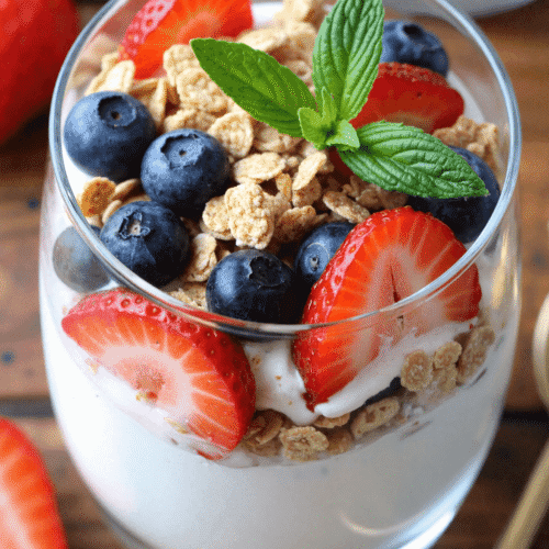Layered high protein Greek yogurt parfait with blueberries, strawberries, granola, and honey drizzle in a tall glass