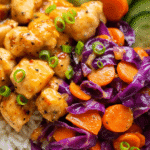 A close-up of juicy Bang Bang Chicken Bites coated in spicy-sweet sauce, surrounded by sautéed carrots and purple cabbage on a bed of white rice.
