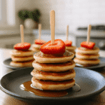 A top-view photograph of a stack of four mini pancakes topped with sliced strawberry and maple syrup, served on a dark ceramic plate in a modern kitchen setting.