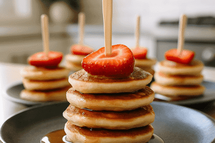 A top-view photograph of a stack of four mini pancakes topped with sliced strawberry and maple syrup, served on a dark ceramic plate in a modern kitchen setting.