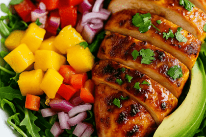 Angled close-up of Sweet Chili Chicken and Mango Salad in a white bowl with grilled chicken glazed in chili sauce, surrounded by mango, avocado, red onion, red bell pepper, and fresh arugula on a wooden background.
