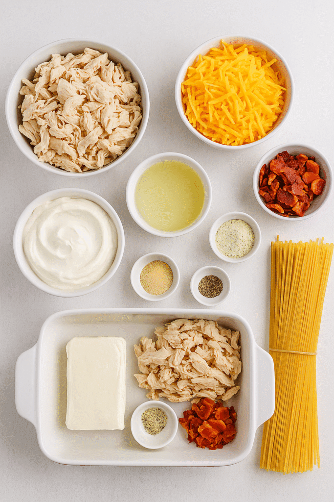 A clean flatlay of crack chicken spaghetti bake ingredients including shredded chicken, cream cheese, sour cream, bacon, ranch mix, cheddar, seasonings, and spaghetti arranged neatly in bowls and a baking dish.