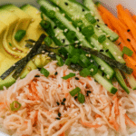 Close-up of a California Roll Sushi Bowl with avocado slices, shredded crab, julienned cucumber and carrots, nori strips, green onions, and sesame seeds over sushi rice.