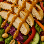 Close-up image of Thai chicken salad highlighting juicy grilled chicken slices, crunchy red cabbage, carrots, cucumbers, and peanuts, all coated in a rich peanut dressing with visible sesame seeds and crisp textures