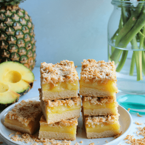 Tropical pineapple coconut bars stacked on a ceramic plate with pineapple slices and toasted coconut – recipe by readyprepmeals