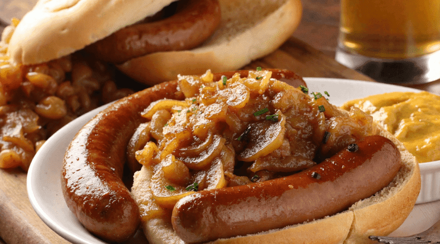 Beer-braised bratwursts served in a rustic dish, topped with caramelized lentils and onions, with mustard and crusty bread on the side.