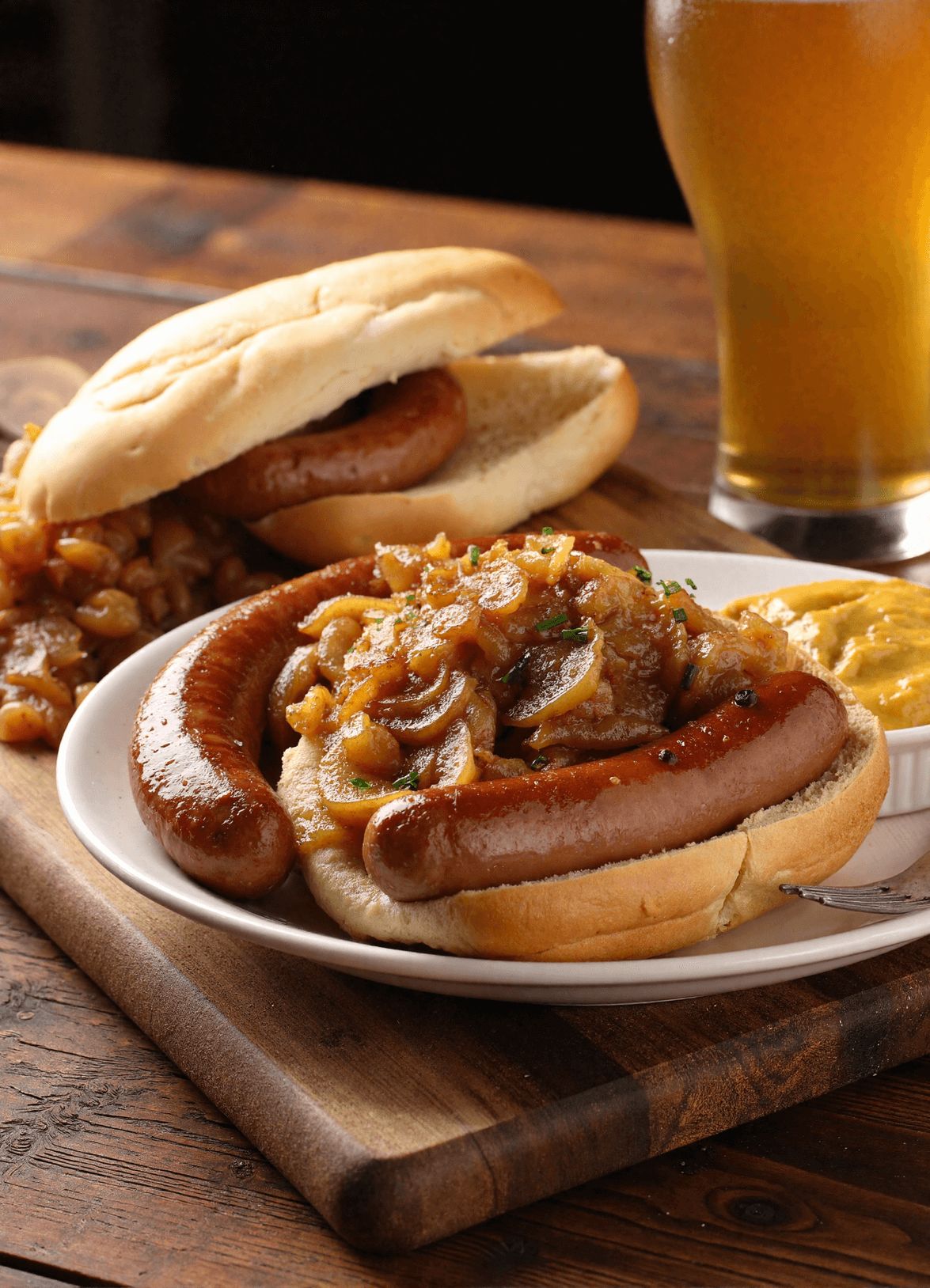 Beer-braised bratwursts served in a rustic dish, topped with caramelized lentils and onions, with mustard and crusty bread on the side.