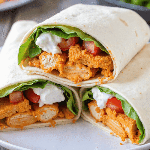 Three buffalo chicken wraps filled with crispy chicken tenders, lettuce, tomato, and blue cheese dressing stacked on a white plate