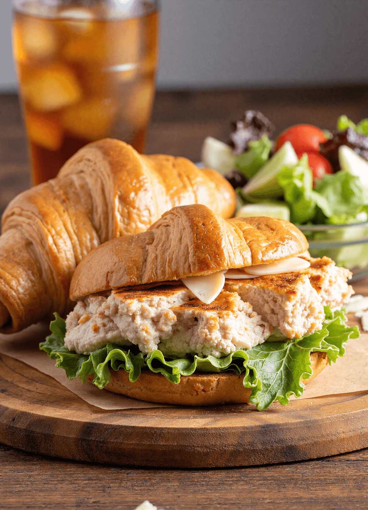 Chicken Caesar Croissant Sandwich on a wooden board with fresh lettuce and iced tea in the background