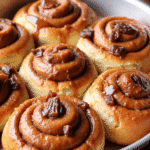 Freshly baked cinnamon date sticky buns in a round baking pan, featuring golden-brown swirls filled with date paste and cinnamon, topped with a glossy glaze and chopped dates, set on a wooden table in a cozy kitchen.