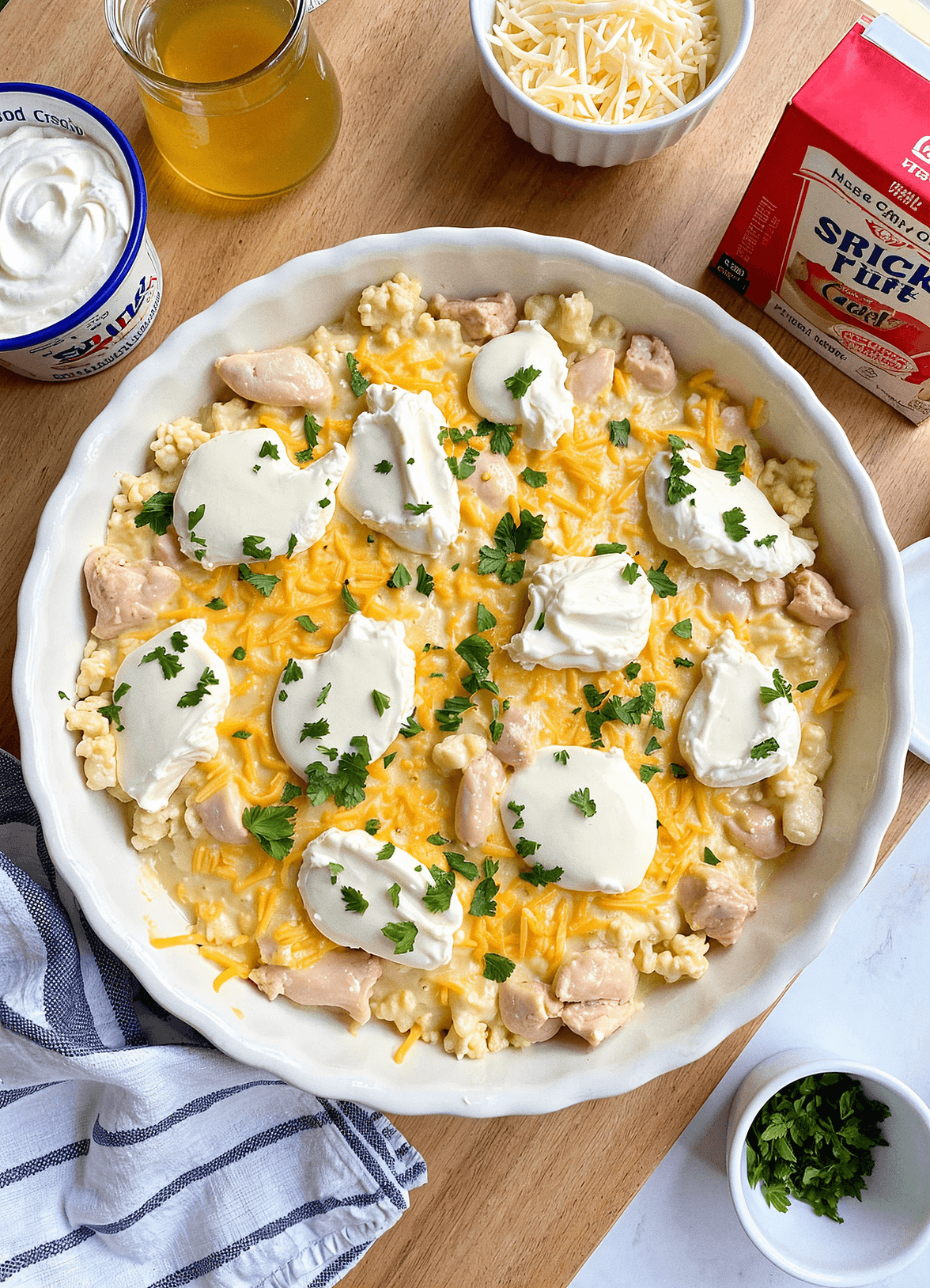 A homemade creamy chicken casserole in a white baking dish, topped with golden breadcrumbs, surrounded by fresh ingredients like sour cream, broth, and shredded cheese, set on a wooden countertop in a bright kitchen.