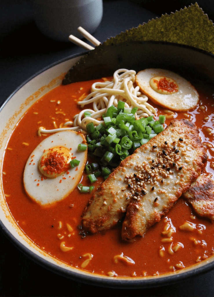 Bowl of fiery chicken ramen with creamy garlic sauce, topped with seared chicken breast, soft-boiled eggs, scallions, and ramen noodles in spicy red broth