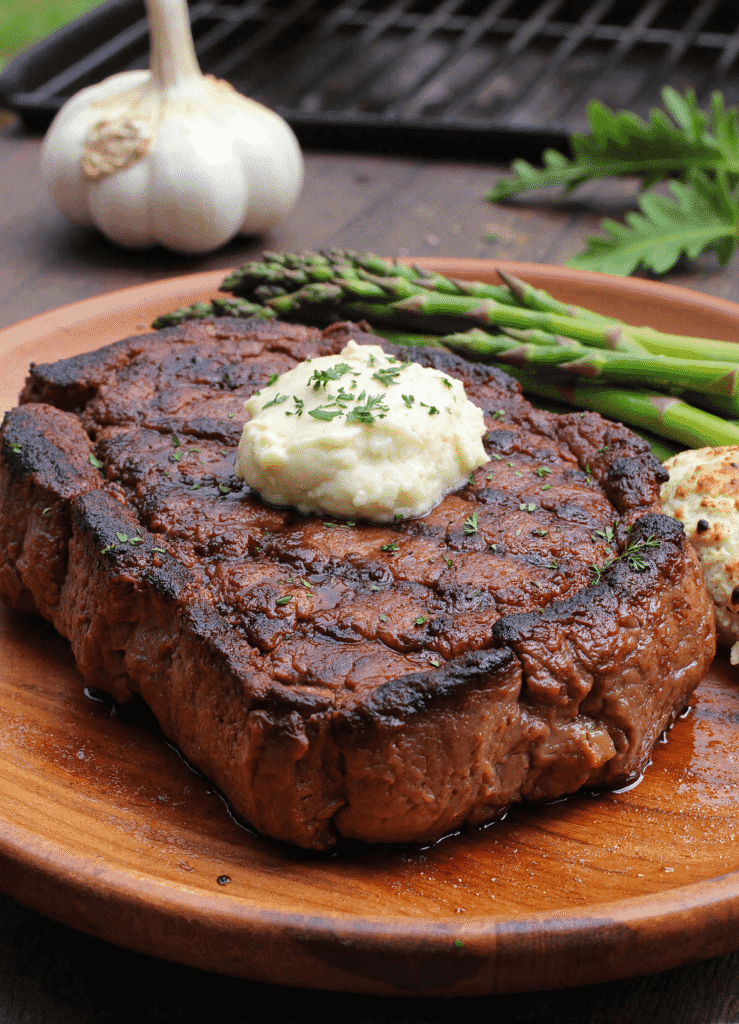 Grilled ribeye steak with a dollop of garlic herb butter served with asparagus and grilled garlic on a wooden plate.