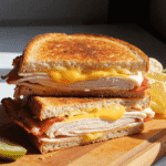 Golden brown grilled turkey bacon melt sandwich with cheddar cheese, stacked on a wooden board and served with pickles and potato chips