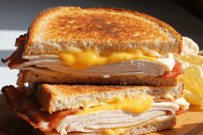 Golden brown grilled turkey bacon melt sandwich with cheddar cheese, stacked on a wooden board and served with pickles and potato chips