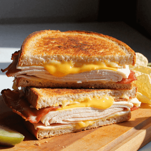 Golden brown grilled turkey bacon melt sandwich with cheddar cheese, stacked on a wooden board and served with pickles and potato chips