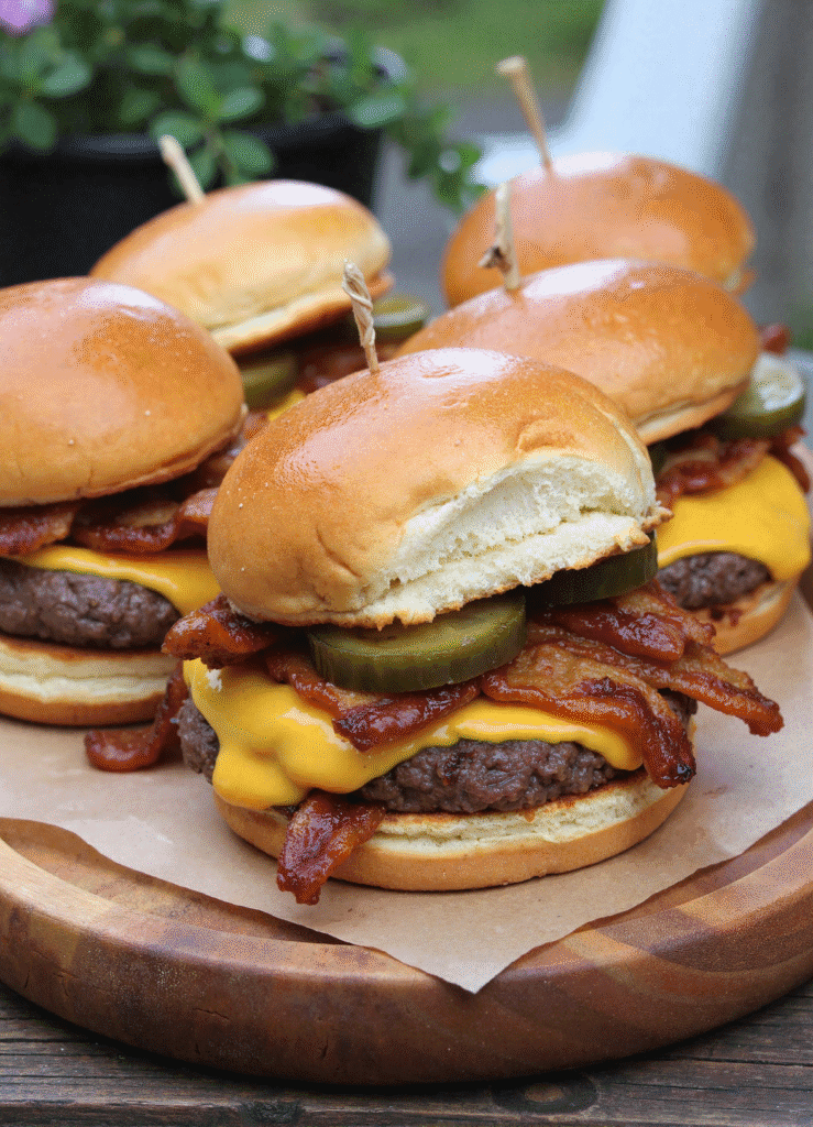 Loaded cheeseburger sliders with cheddar, bacon, and pickles on toasted buns, served on a rustic wooden platter.