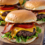 Close-up of loaded cheeseburger sliders with cheddar, crispy bacon, lettuce, and tomato on toasted buns.