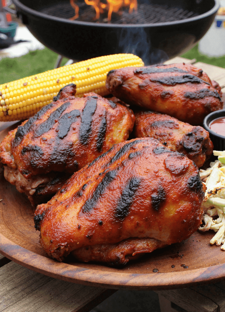 Grilled smoky BBQ chicken thighs with visible char marks, served with corn on the cob and cabbage slaw.