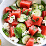 Watermelon feta salad with mint and cucumber served in a white ceramic bowl on a rustic wood table, garnished with fresh herbs and lime wedges