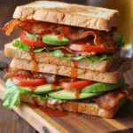 Stacked spicy BLT avocado sandwich dripping with chipotle mayo, layered with crispy bacon, avocado slices, lettuce, and tomato on toasted sandwich bread.