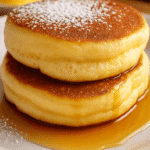 A close-up digital photograph captures a stack of two fluffy soufflé pancakes with powdered sugar and maple syrup on a white plate, highlighting their airy texture.