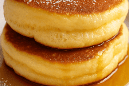 A close-up digital photograph captures a stack of two fluffy soufflé pancakes with powdered sugar and maple syrup on a white plate, highlighting their airy texture.
