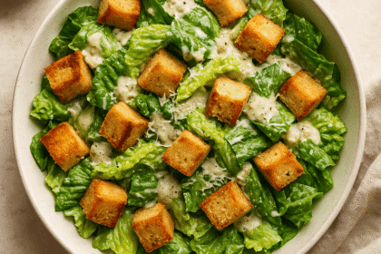 A top view of a vegan Caesar salad in a large white bowl, featuring crisp romaine lettuce, golden croutons, and creamy aquafaba-based dressing, sprinkled with vegan Parmesan and fresh cracked pepper.