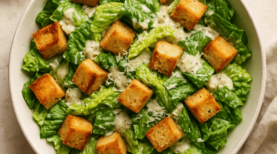 A top view of a vegan Caesar salad in a large white bowl, featuring crisp romaine lettuce, golden croutons, and creamy aquafaba-based dressing, sprinkled with vegan Parmesan and fresh cracked pepper.