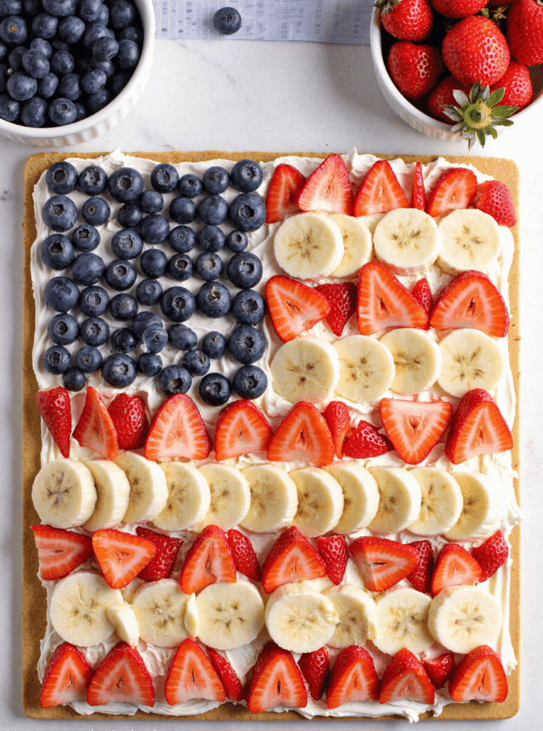 Close-up of American Flag Fruit Pizza with sugar cookie crust, whipped cream cheese frosting, and rows of strawberries, blueberries, and bananas in a patriotic red, white, and blue pattern.