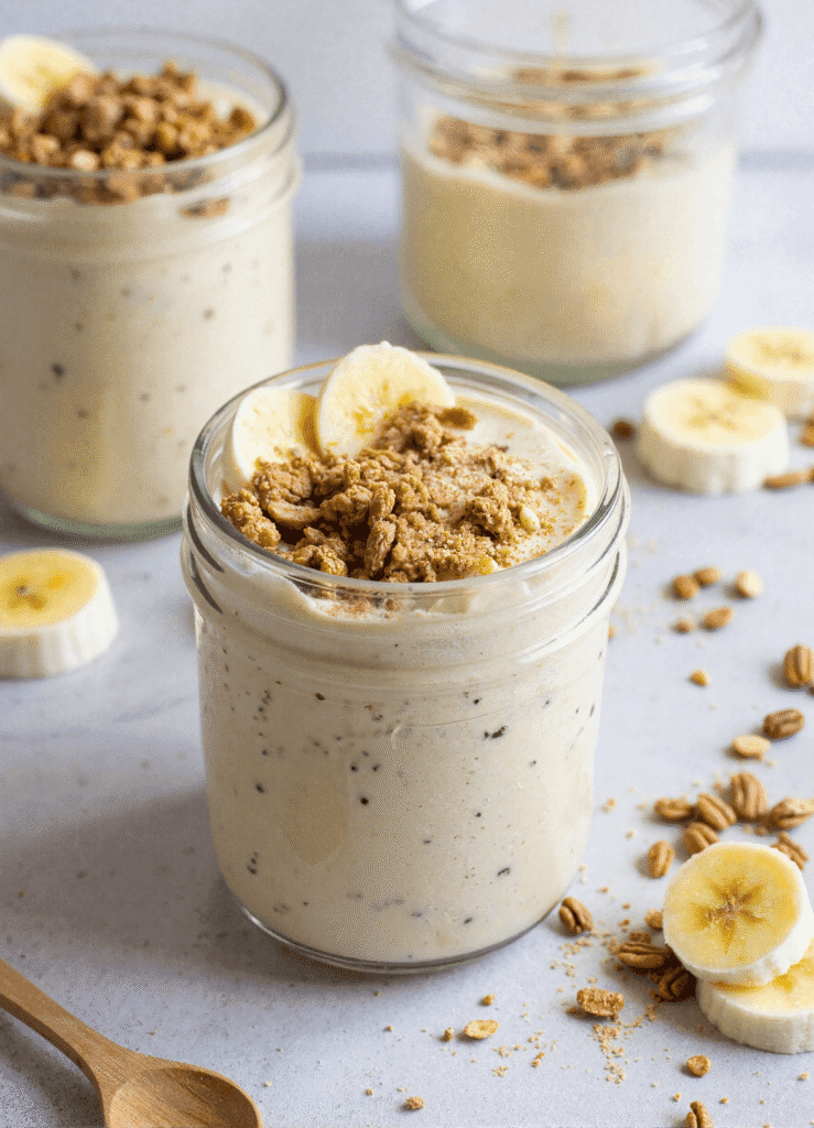 Jar of banana protein pudding topped with banana slices and granola, surrounded by oats and fresh banana slices