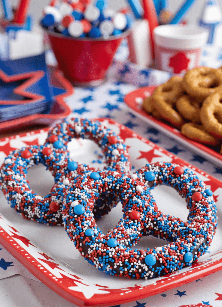 Chocolate-covered pretzel rings coated in red, white, and blue sprinkles on a patriotic plate for a festive 4th of July dessert.