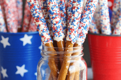 White chocolate-dipped pretzel rods decorated with red and blue sprinkles standing upright in a mason jar for a 4th of July party.