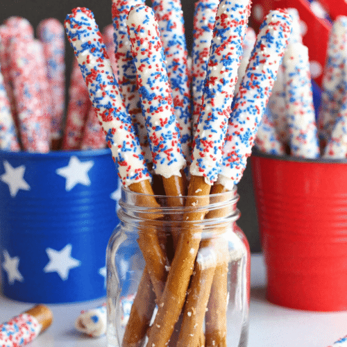 White chocolate-dipped pretzel rods decorated with red and blue sprinkles standing upright in a mason jar for a 4th of July party.