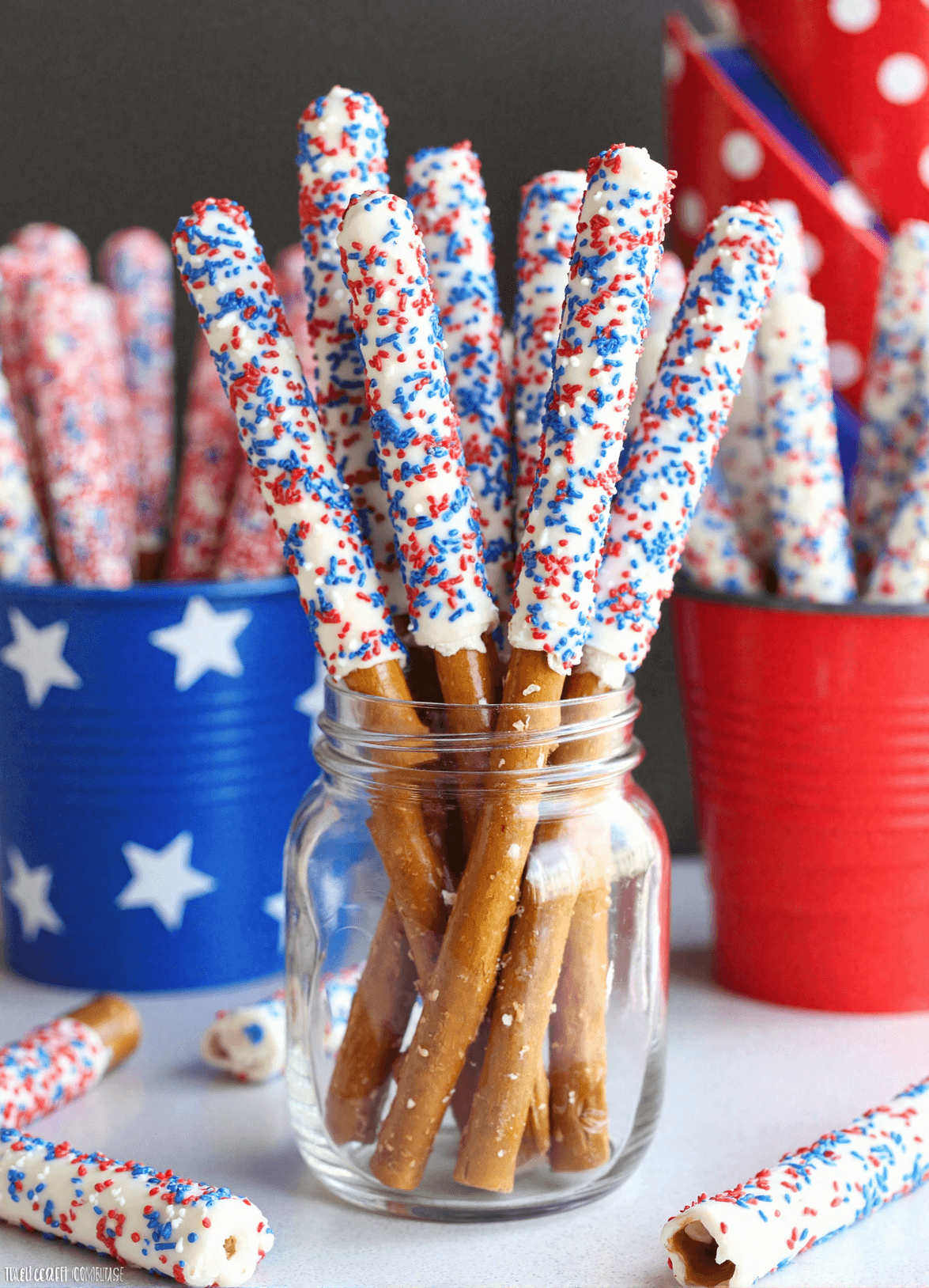 White chocolate-dipped pretzel rods decorated with red and blue sprinkles standing upright in a mason jar for a 4th of July party.