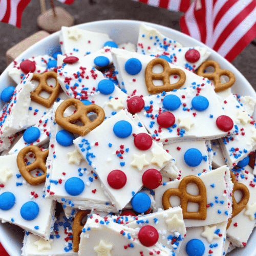 Bowl of Fourth of July pretzel bark topped with white chocolate, red and blue M&Ms, star sprinkles, and whole mini pretzels with American flags in the background.