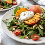 Grilled peach and burrata salad on a white plate with arugula, cherry tomatoes, almonds, and balsamic glaze