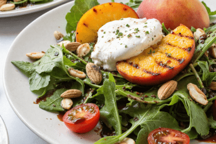 Grilled peach and burrata salad on a white plate with arugula, cherry tomatoes, almonds, and balsamic glaze