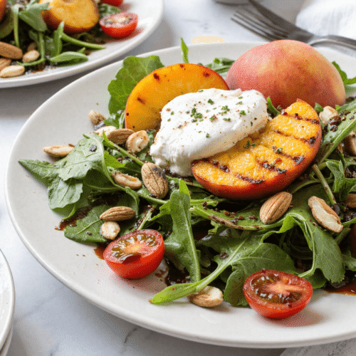 Grilled peach and burrata salad on a white plate with arugula, cherry tomatoes, almonds, and balsamic glaze