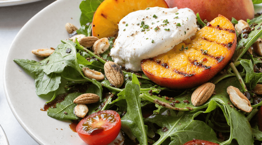 Grilled peach and burrata salad on a white plate with arugula, cherry tomatoes, almonds, and balsamic glaze