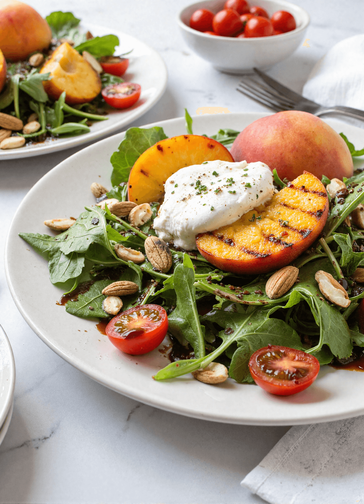 Grilled peach and burrata salad on a white plate with arugula, cherry tomatoes, almonds, and balsamic glaze