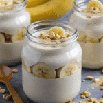 Creamy high protein banana pudding in glass jars layered with banana slices and granola, served on a gray stone background