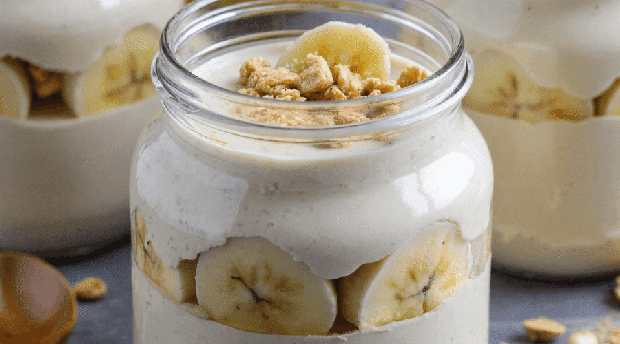 Creamy high protein banana pudding in glass jars layered with banana slices and granola, served on a gray stone background