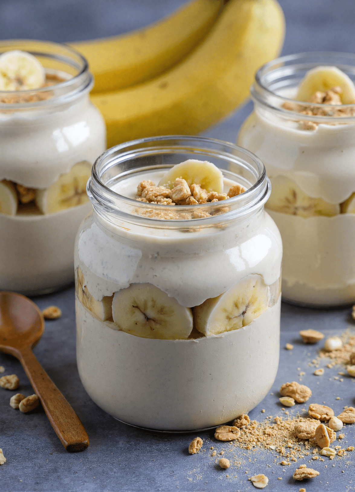 Creamy high protein banana pudding in glass jars layered with banana slices and granola, served on a gray stone background
