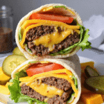 Close-up of a high protein cheeseburger wrap with melted cheddar, ground beef, lettuce, and tomato stacked on a plate with pickles.