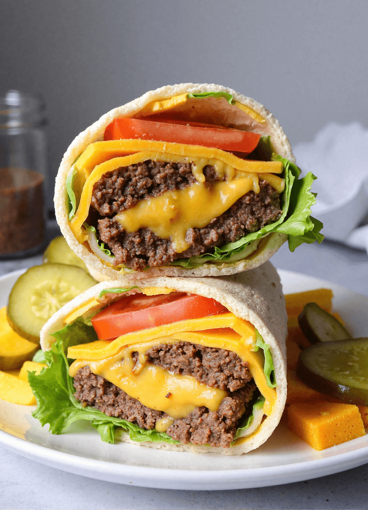 Close-up of a high protein cheeseburger wrap with melted cheddar, ground beef, lettuce, and tomato stacked on a plate with pickles.