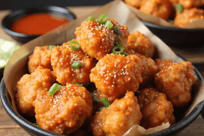 Bowl of Korean chicken bites glazed in spicy gochujang sauce, garnished with sesame seeds and green onions, with a dipping sauce in the background.