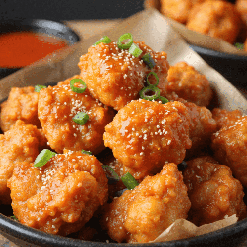 Bowl of Korean chicken bites glazed in spicy gochujang sauce, garnished with sesame seeds and green onions, with a dipping sauce in the background.